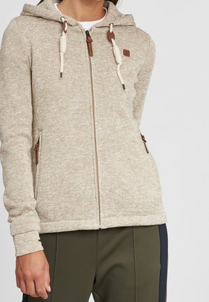 Femme portant un pull à capuche beige avec fermeture éclair et cordons, ainsi qu’un pantalon vert olive, debout devant un fond clair uni.
