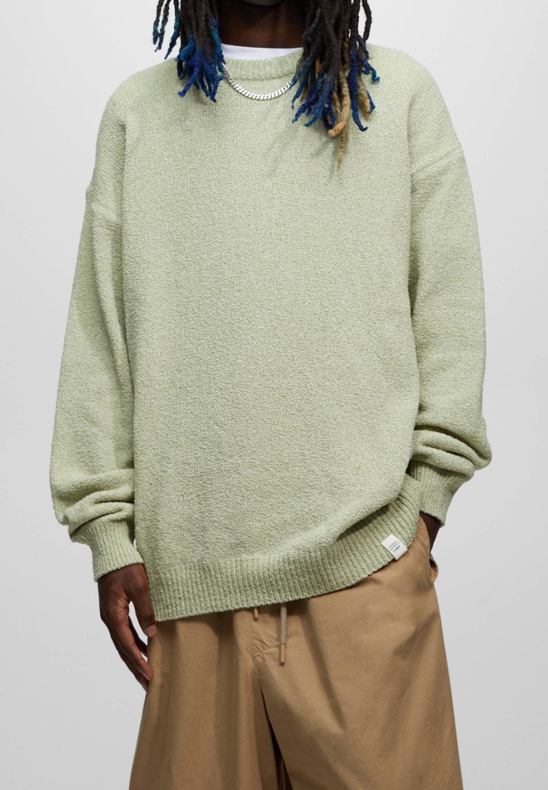 Homme portant un pull vert clair oversize et un pantalon beige, une main dans la poche, debout devant un fond uni.