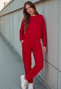 Červená mikina a odpovídající jogger kalhoty z měkké tkaniny, s volným střihem. Bílé tenisky doplňují outfit, před šedou zdí.