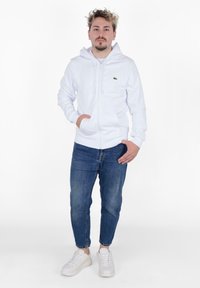 Felpa bianca con zip e tasche a canguro, abbinata a jeans blu e sneakers bianche. La felpa presenta un piccolo logo verde sul petto.