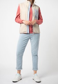 Fleece vesta v krémové barvě s světle modrými akcenty, se zipovými kapsami, v kombinaci s růžovou košilí s dlouhým rukávem a světle modrými kordovými kalhotami.