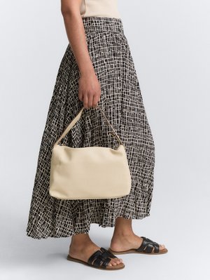 Bolso texturizado beige con forma redondeada, sostenido por una persona que lleva una falda larga de estampado blanco y negro y sandalias negras.