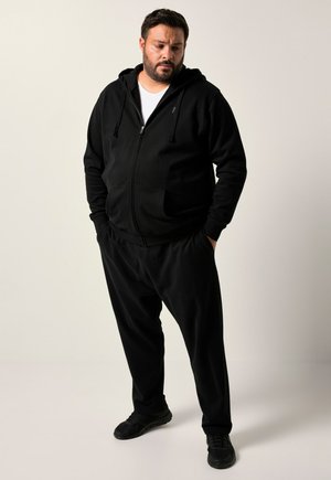 Mand med skæg iført sort hoodie, sorte bukser og sorte sneakers, stående med hænderne i lommerne mod en ensfarvet lys baggrund.