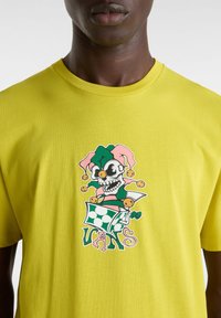 Camiseta amarilla de manga corta con un gráfico central de una calavera con gorro de bufón y acentos coloridos, y el texto "VANS" en una fuente estilizada.