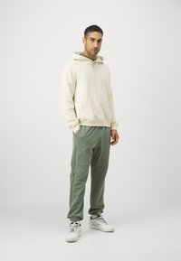 Light beige hoodie with a front pocket, paired with olive green joggers featuring ribbed cuffs. White sneakers complete the outfit.