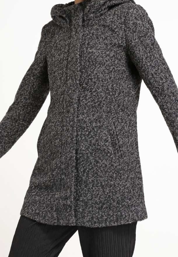Femme portant un manteau en laine texturé gris foncé avec capuche et fermeture frontale cachée, associé à un pantalon plissé noir.