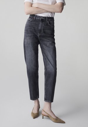 Osoba ubrana w wyblakłe czarne spodnie dżinsowe z wysokim stanem i beżowe zamszowe sandały na obcasie z odkrytą piętą i zakończone szpiczastym noskiem, z założonymi skrzyżowanymi ramionami, na jednolitym jasnym tle.