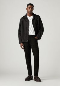 Tânăr stând cu mâna în buzunar, purtând o jachetă neagră cu nasturi, un tricou alb, pantaloni negri și pantofi maro închis pe un fundal simplu.