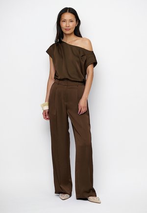 Vrouw draagt een bruine one-shoulder top en bijpassende wijde broek, beige puntige schoenen en witte armbanden, staand tegen een witte achtergrond.