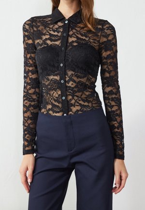 Mujer vistiendo una blusa de encaje floral negra de manga larga con botones sobre un bandeau negro, combinada con pantalones de talle alto color azul marino oscuro.