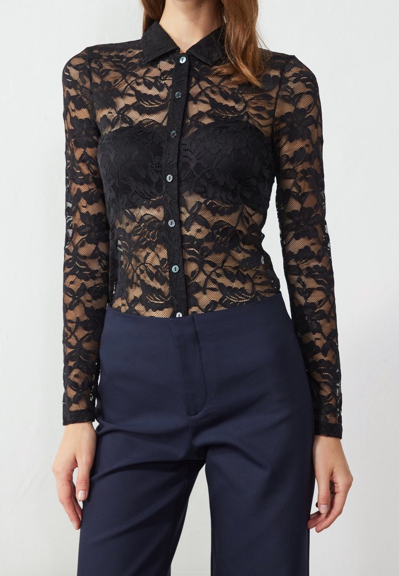 Femme portant un haut noir à manches longues en dentelle florale avec des boutons, par-dessus un bandeau noir, associé à un pantalon taille haute bleu marine foncé.