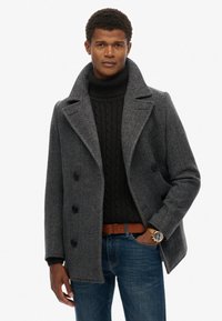 Uomo con cappotto doppiopetto grigio sopra un dolcevita nero a trecce, jeans blu e cintura marrone, con una mano in tasca.