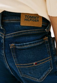 Džínsy v tmavomodrej farbe s oranžovým prešívaním. Obsahujú hnedú koženú etiketu so značkou "TOMMY HILFIGER" a malým logom na zadnej vrecku.