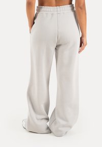 Femme portant un pantalon de survêtement gris clair ample et des baskets blanches, debout, les mains partiellement dans les poches, vue de dos.