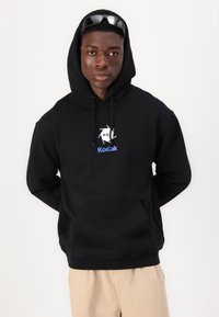 Black hoodie with a drawstring, featuring a white and blue Kodak logo. Soft fabric with a front pocket. Model stands against a white background.