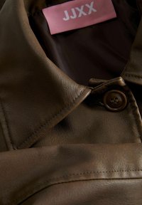 Giacca di pelle marrone con colletto a punte, dotata di una chiusura a un bottone, dal tocco morbido e un'etichetta rosa con la scritta "JJXX" all'interno.