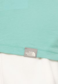 Jasno niebieska bawełniana koszulka z prostym krojem, z szarym metką z napisem "The North Face" u dołu. Gładka tekstura, casualowy design.