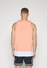 Persik mesh tank topp med hvit bunnkant, har en avslappet passform og perforert tekstur. Brukt med svarte treningsshorts.