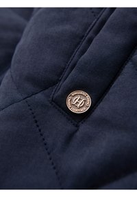 Tessuto trapuntato blu scuro con un bottone rotondo in bronzo recante la scritta "Ombre Heritage" e dettagli di cucitura intricati.