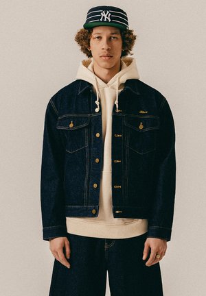 Junger Mann mit lockigem Haar trägt eine dunkle Jeansjacke über einem beigen Hoodie und eine schwarz-weiß gestreifte Baseballkappe.