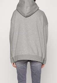 Felpa grigia oversize in morbido mix di cotone, presenta un grande cappuccio, spalle cadenti e polsini a coste, con un orlo posteriore curvato verso l'alto.