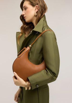 Žena v olivovém kabátu nese hnědou koženou tašku ve tvaru půlměsíce s pleteným popruhem a nosí zlaté kruhové náušnice a náramky.