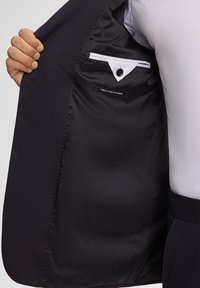 Zwart blazer interieur met gladde stof, rode stiksels, zak met knoopsluiting en label "Finest Stretch Fabric".