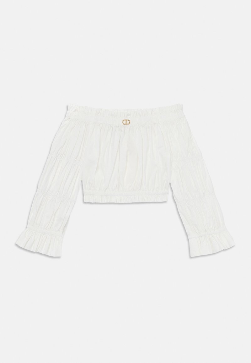 Fehér, vállnélküli blúz, gyűrött textúrával, háromnegyedes ujjal és fodros mandzsettával. A nyakán egy kis arany díszítés található.