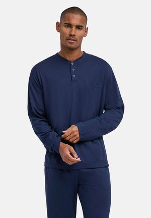 Mann trägt ein navyblaues langärmeliges Henley-Shirt mit drei Knöpfen und passende Hosen vor einem schlichten Hintergrund.