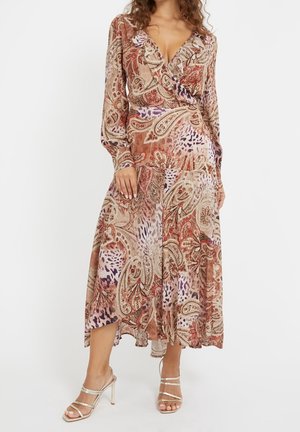 Kobieta w sukience midi z długimi rękawami i dekoltem w serek, w wzór paisley w kolorach czerwonym, beżowym i fioletowym, w połączeniu z beżowymi sandałami na paskach.