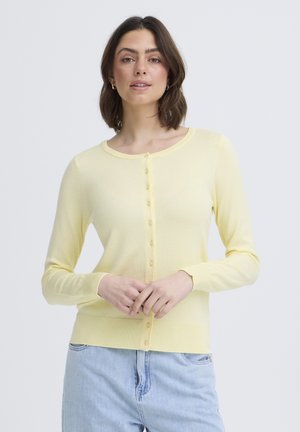 Žena s hnedými vlasmi po plecia, oblečená v bledozelený zapínaný kardigán a svetlomodré džínsy, stojaca so založenými rukami.