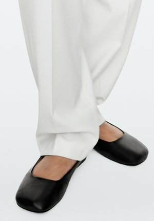 Pieds portant des ballerines noires à bout carré associées à un pantalon blanc ample sur un fond uni.