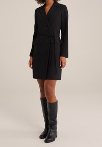 Svart skräddarsydd klänning med långa ärmar och knytning i midjan. Har två framfickor och slagkrage. Kombineras med knähöga svarta läderstövlar.