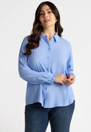 Camicia azzurra chiaro con bottoni e maniche lunghe, dalla texture liscia e con polsini elasticizzati, abbinata a jeans blu scuro.