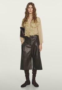 Tan rynget bluse parret med brune læder culottes, der har brede ben og lommer. Brune knæhøje, rynkede støvler fuldender looket.