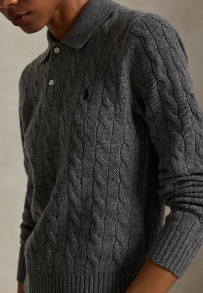 Henkilö, jolla on yllään tummanharmaa palmikkoneulepaita, jossa on kaulus ja napit, ja rinnassa pieni kirjailtu logo.