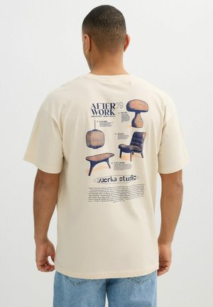 Hombre con camiseta beige con ilustraciones de muebles y texto en la parte trasera, incluyendo diseños de silla, mesa, lámpara y cojín.