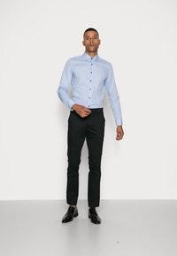Lichtblauwe blouse met kraag en verticale strepen, geknoopt aan de voorkant, gecombineerd met zwarte slim-fit broek en zwarte schoenen op een witte achtergrond.
