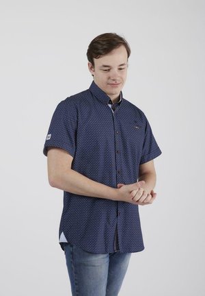 Chemise bleue marine à manches courtes avec des pois blancs, un col et des motifs contrastants ; associée à un jean bleu clair.
