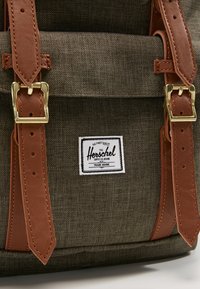 Mochila de tela verde con correas de cuero marrón y hebillas doradas, que presenta una etiqueta de la marca Herschel Supply Co. en blanco.