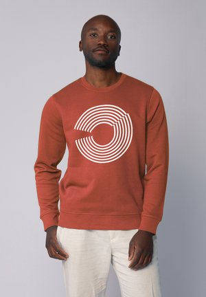 Roestkleurige sweatshirt met een grote witte geconcentreerde cirkelgrafiek aan de voorkant, met een ronde halslijn en ribgebreide manchetten.