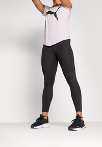 Leggings deportivos negros combinados con una camiseta de manga corta rosa claro con el logo negro de Puma. Los zapatos son negros con detalles en verde y blanco.