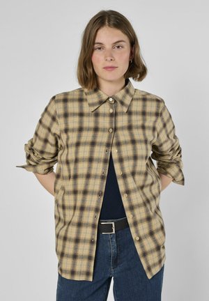 Kariertes Hemd mit Knopfleiste in Beige- und Schwarztönen, mit klassischem Kragen, langen Ärmeln und lockerem Schnitt, kombiniert mit dunkler Jeans und einem Gürtel.