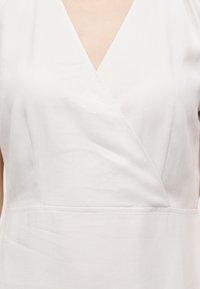 Λευκό τοπ με V-λαιμόκοψη τύπου wrap και φαρδιές τιράντες, κατασκευασμένο από υφή υφάσματος, εμφανιζόμενο από το στήθος έως τη μέση.