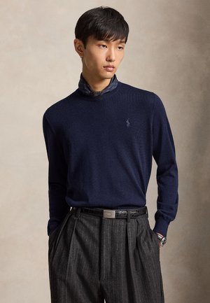 Jeune homme portant un pull bleu marine par-dessus une chemise à motifs, un pantalon gris à fines rayures, une ceinture noire et une montre-bracelet, debout les mains dans les poches.