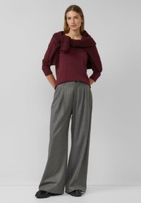 Bordeaux lange mouw top gecombineerd met grijze streepjesspijkerbroek met wijde pijpen, ceintuur om de taille. Trui casual over de schouders gedragen. Zwarte schoenen.