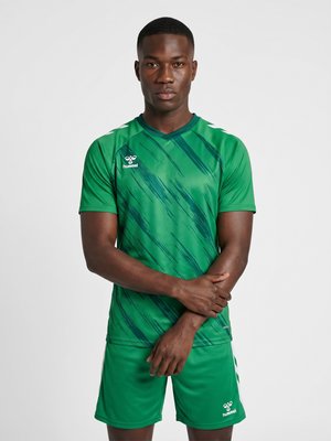 Mann trägt grünes Sporttrikot und Shorts mit dunklen diagonalen Streifen, steht mit verschränkten Armen vor einfarbigem Hintergrund.