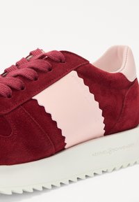 Baskets en suède rouge avec un accent en cuir rose, présentant un design festonné, des lacets ronds et une semelle blanche crantée. Logo de la marque embossé.