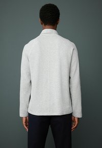 Lys grå sweater med en struktureret overflade, raglanærmer og en flad krave, båret med navy bukser, set fra ryggen mod en mørk baggrund.