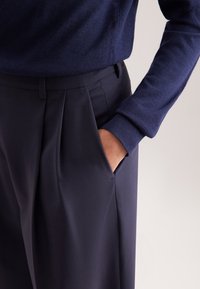 Persoon in donkerblauwe geplooide broek en een bijpassende donkerblauwe trui met lange mouwen, met één hand in de zak.